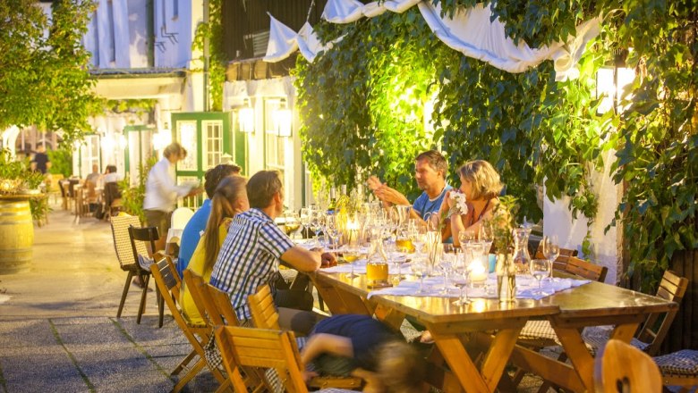 Menschen sitzen an einem langen Tisch im Freien, umgeben von Pflanzen und Lichtern.