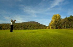 Golfer schl&auml;gt Ball auf einem gr&uuml;nen Golfplatz mit bewaldeten H&uuml;geln im Hintergrund.