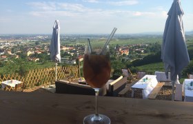 Ein Getränk in einem Weinglas mit Strohhalmen vor einer Landschaft mit Tischen und Sonnenschirmen.