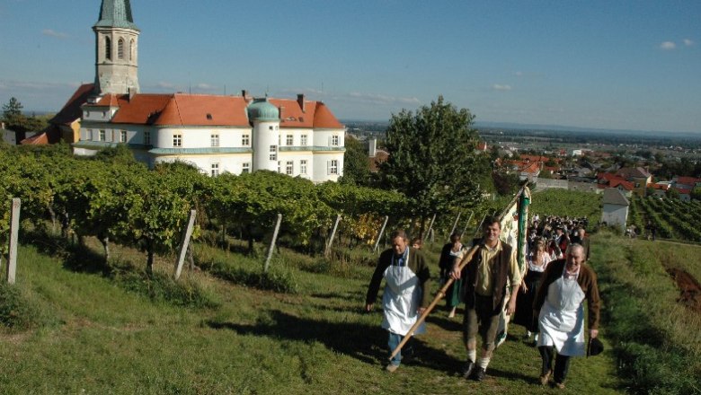 Menschen in traditioneller Kleidung gehen durch Weinberge in Gumpoldskirchen, im Hintergrund eine Kirche und Geb&auml;ude.